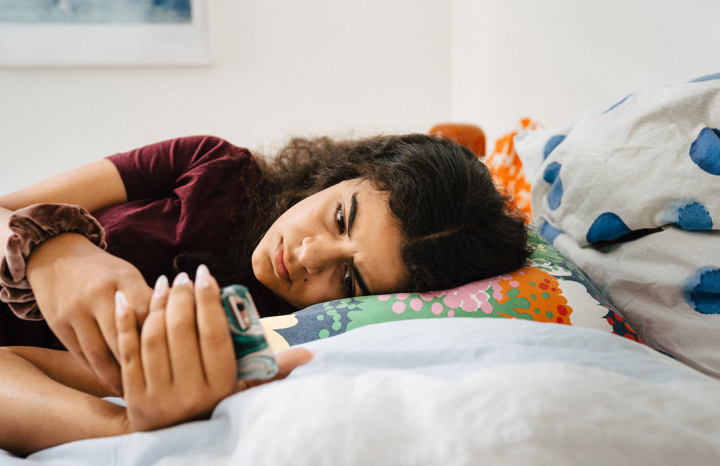 Tjej ligger på sin säng och tittar på telefonen med orolig blick.